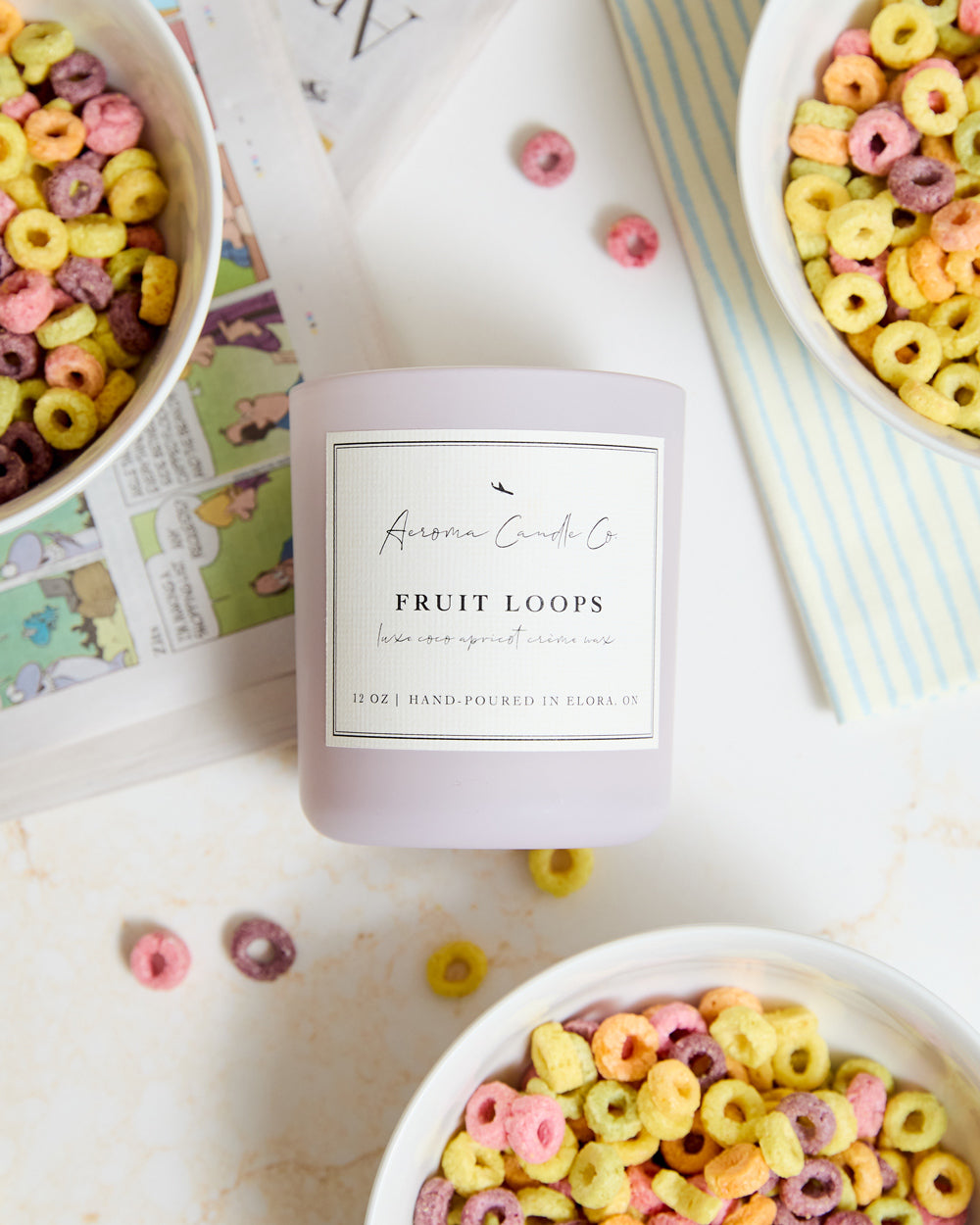 An above-shot of the "Fruit Loops" candle surrounded by bowls of cereal and coloured comics. The candle has a white label and the vessel is light purple in colour.