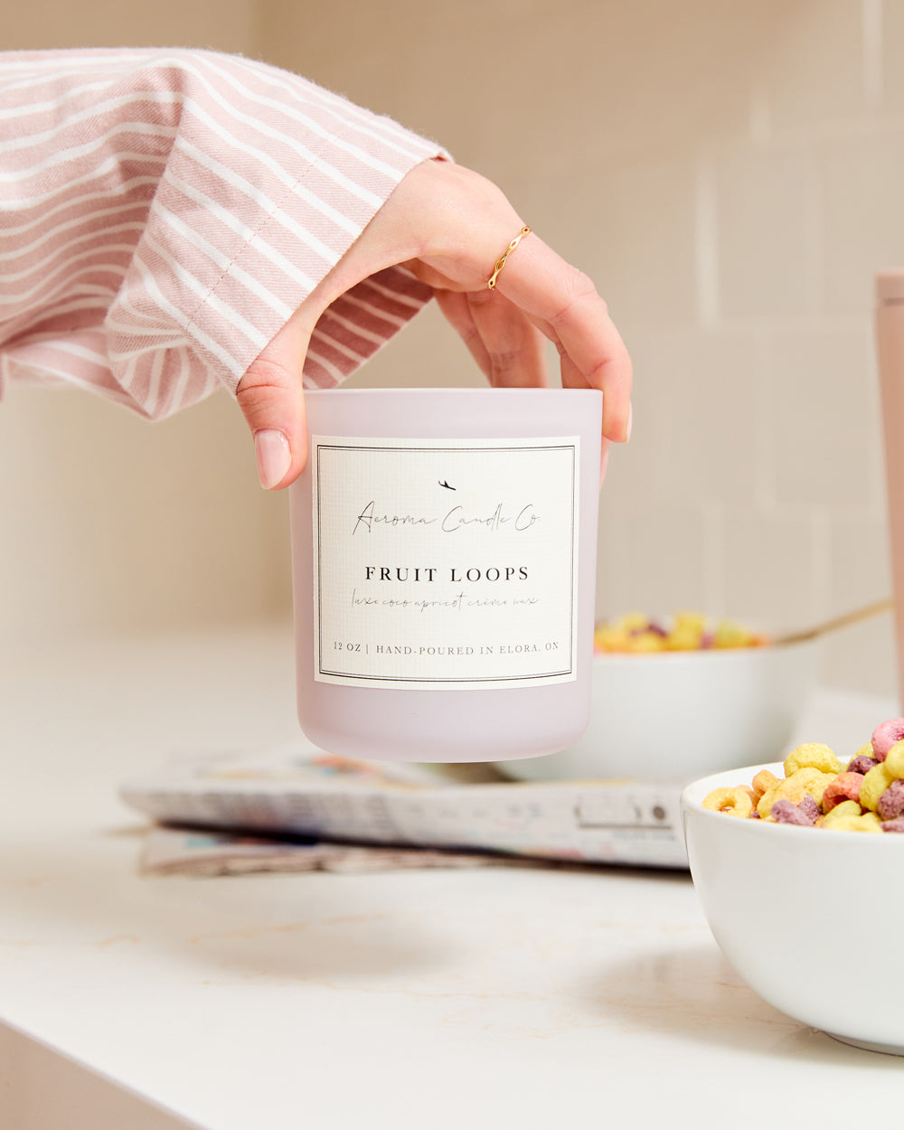 A close up of a hand lifting up the light purple coloured "Fruit Loops" candle on a kitchen countertop surrounded by cereal bowls and comics.