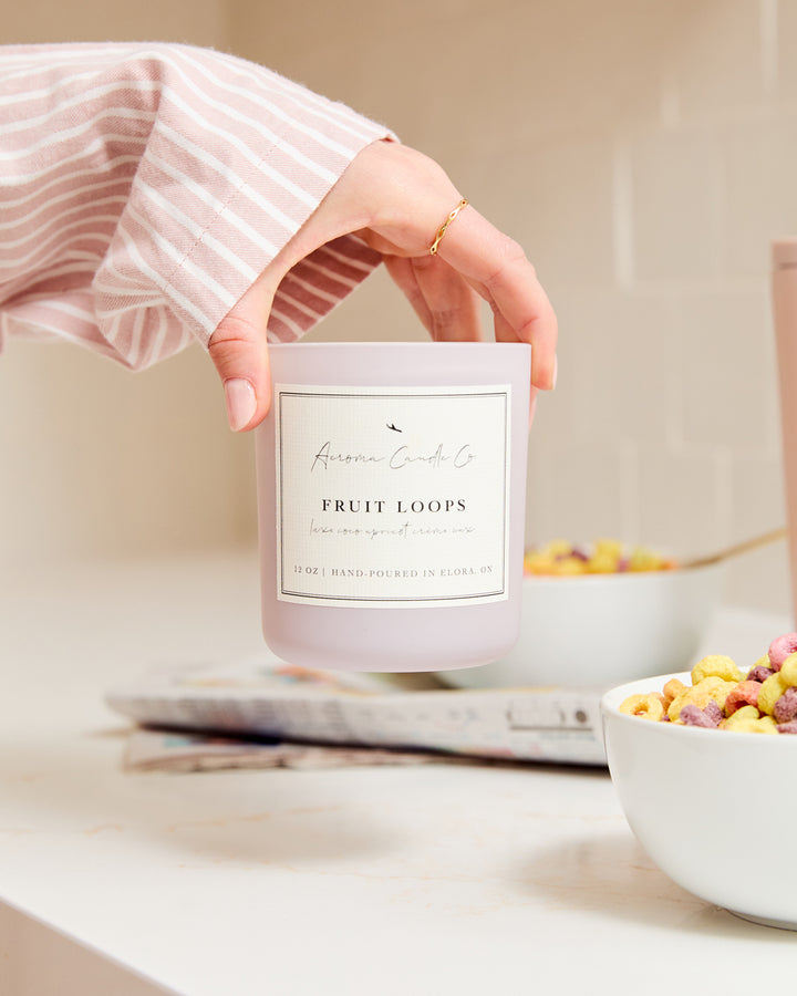 A close up of a hand lifting up the light purple coloured "Fruit Loops" candle on a kitchen countertop surrounded by cereal bowls and comics.