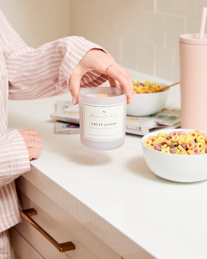 A hand lifting up the light purple coloured "Fruit Loops" candle on a kitchen countertop surrounded by cereal bowls and comics.