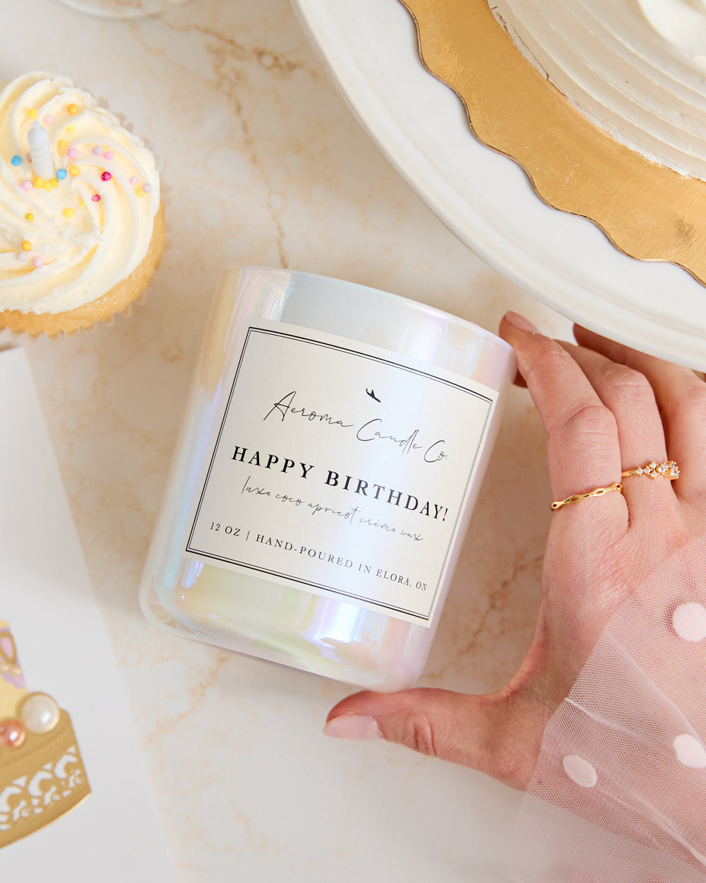 A hand holding the "Happy Birthday" candle over a countertop. Shot at an upward angle, there is a cupcake beside the white iridescent candle vessel.