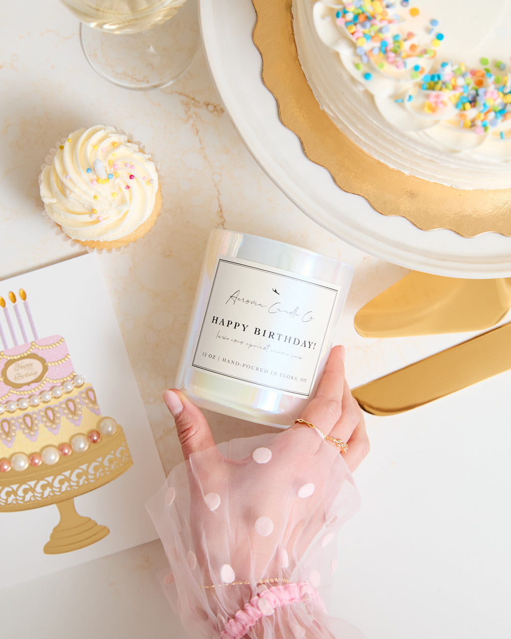 A hand touching the "Happy Birthday" candle over a countertop. Shot at an upward angle, there is a birthday card, cupcake, and full cake surrounding the white iridescent candle vessel.