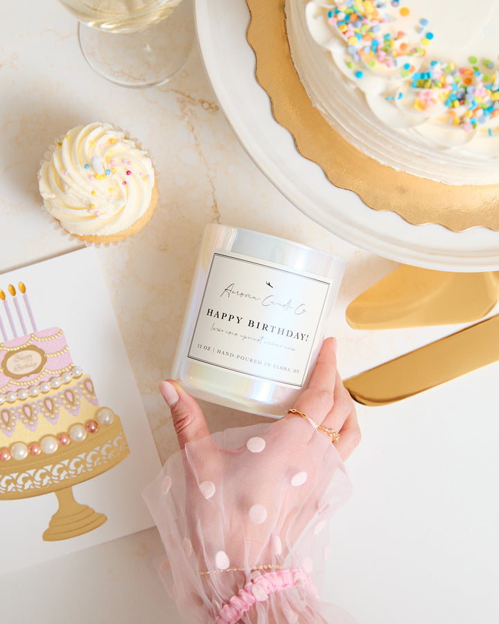 A hand touching the "Happy Birthday" candle over a countertop. Shot at an upward angle, there is a birthday card, cupcake, and full cake surrounding the white iridescent candle vessel.