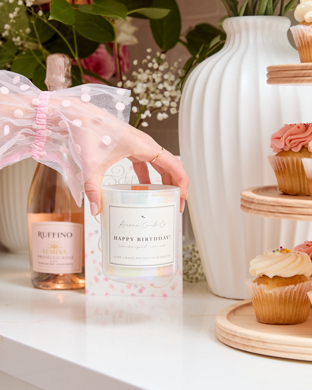 A hand holding the "Happy Birthday" candle from the top over a countertop. The candle vessel is a white iridescent colour. In the background, you can see flowers, cupcakes, and wine.