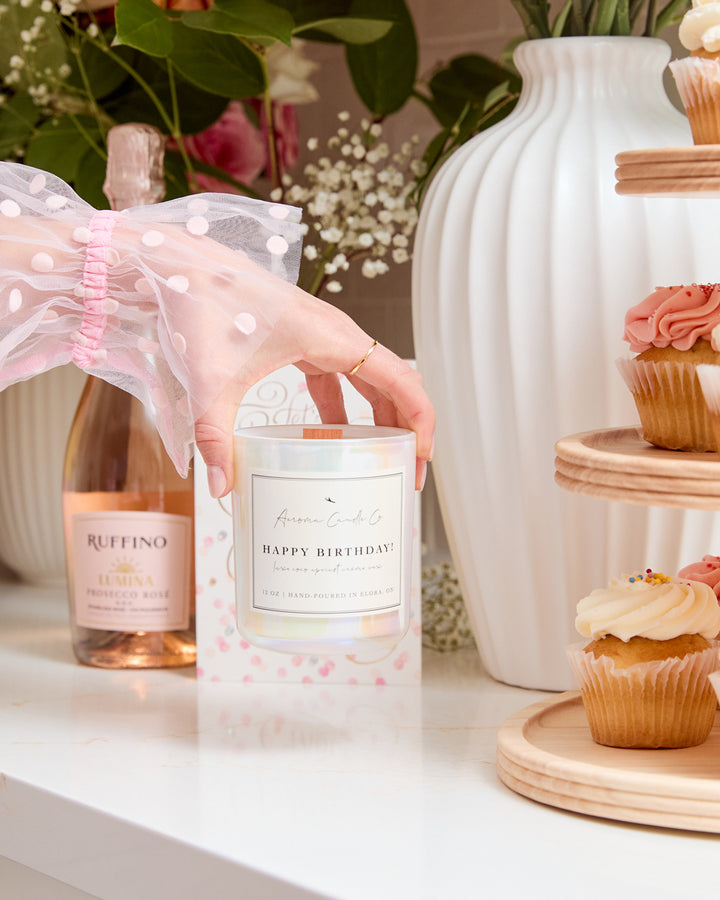 A hand holding the "Happy Birthday" candle from the top over a countertop. The candle vessel is a white iridescent colour. In the background, you can see flowers, cupcakes, and wine.