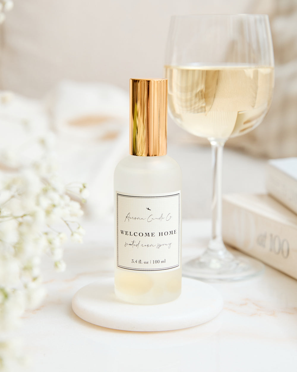 A "Welcome Home" room spray sitting on a coaster beside a glass of white wine in a bright room