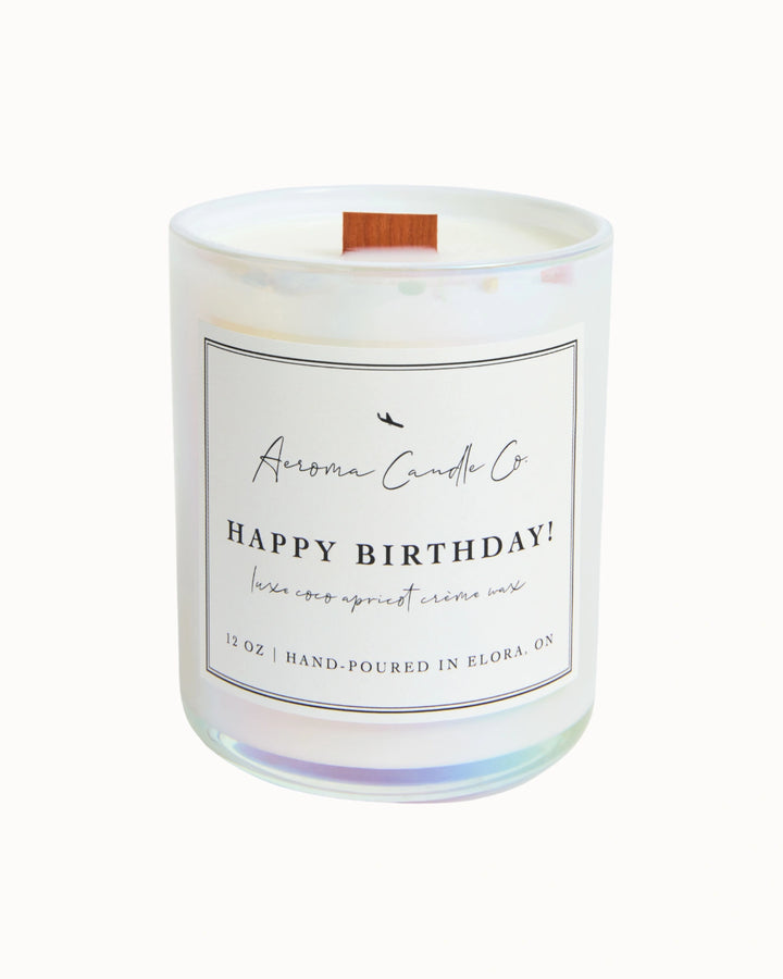 "Happy Birthday" candle in a white iridescent jar. There is a white square label on the candle stating the name of the business, scent, "luxe coco apricot crème wax", size, and "hand-poured in Elora, ON".