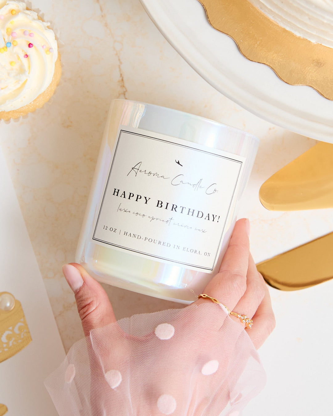Hand holding a 'Happy Birthday' candle with a cake-themed card and pastries in the background