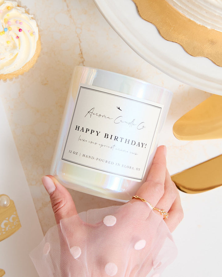 Hand holding a 'Happy Birthday' candle with a cake-themed card and pastries in the background