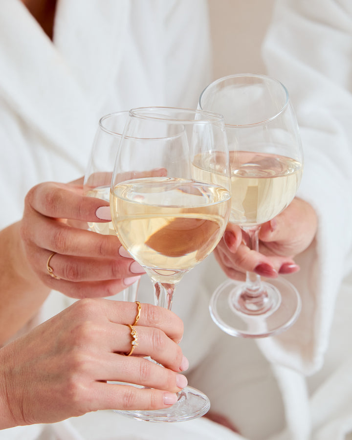 Three women in bathrobes holding glasses of wine together for a cheers in a hotel room.