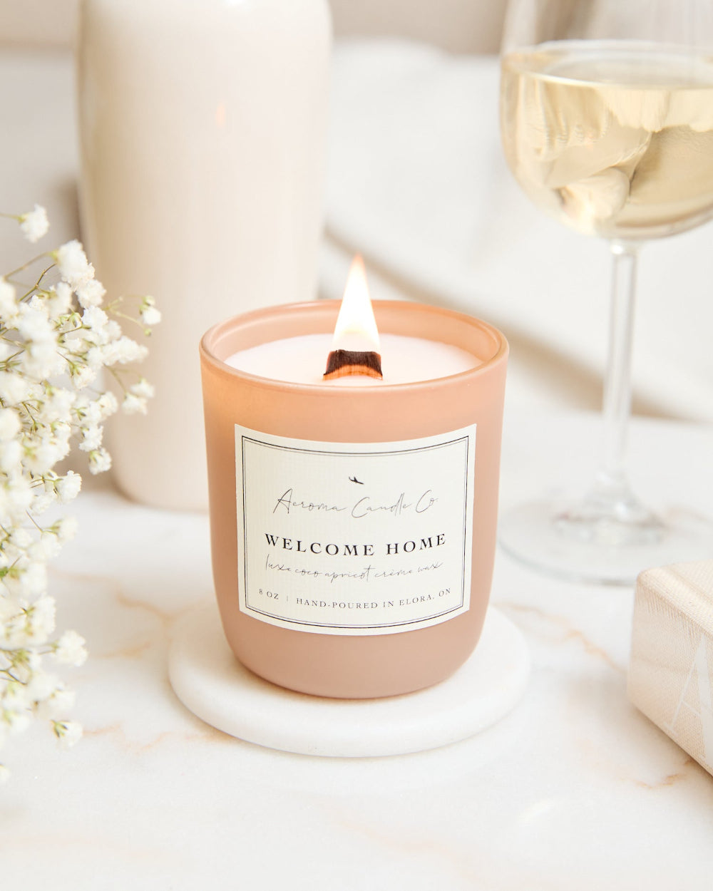 A khaki-colored glass candle on a white marble coaster, surrounded by white baby's breath flowers and a glass of white wine on a light marble surface. The label on the candle reads 'Aeroma Candle Co.'