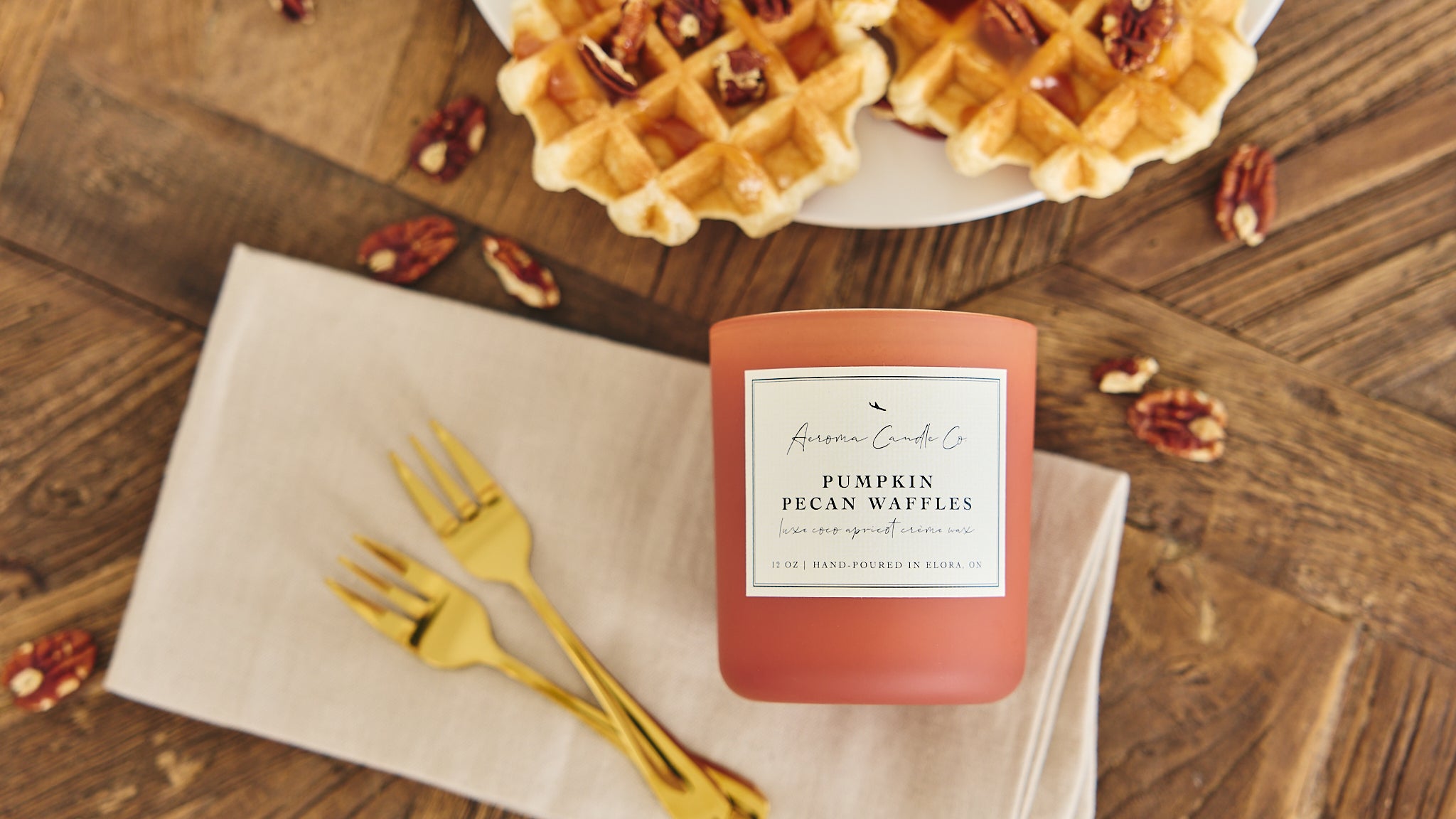 Candle labeled 'Pumpkin Pecan Waffles' on a wooden table with waffles and pecans.