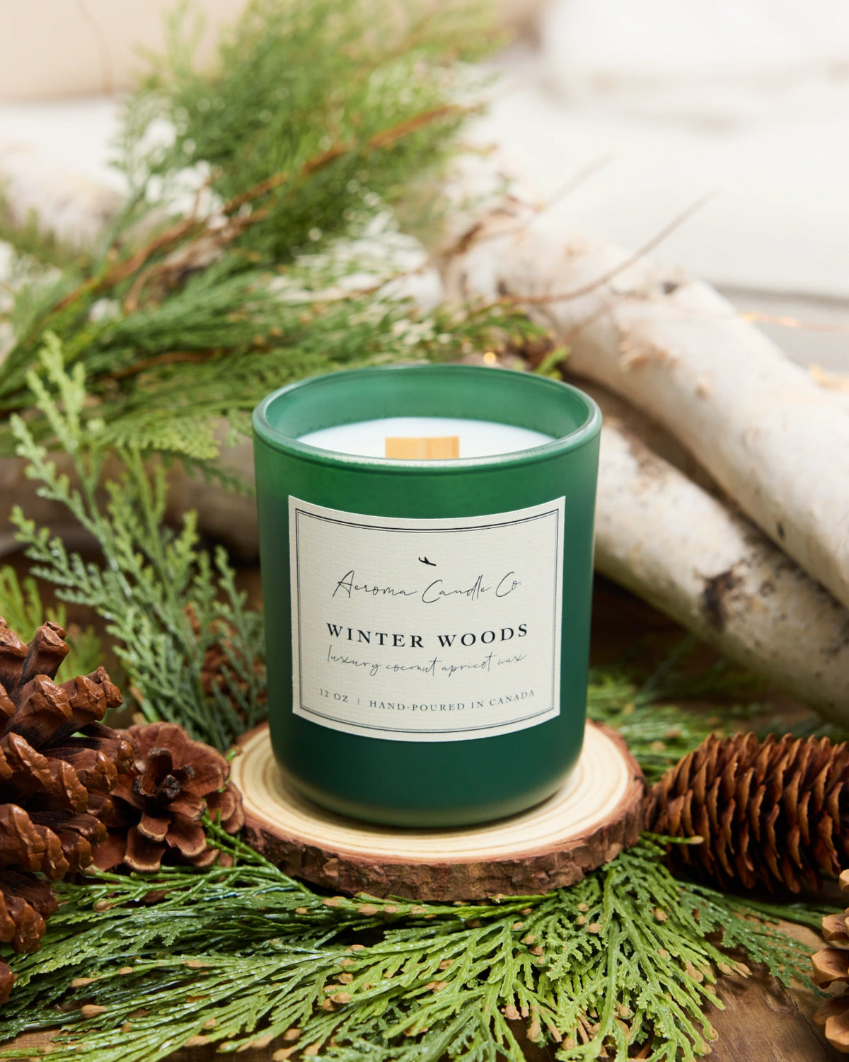Green candle labeled 'Winter Woods' surrounded by greenery and pinecones on a wooden surface