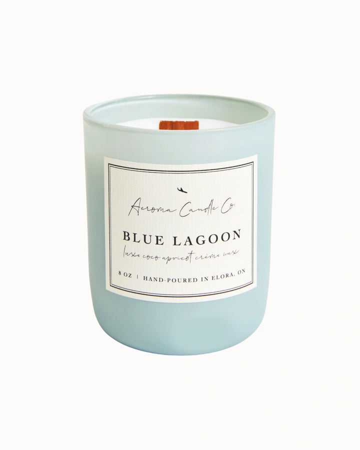 "Blue Lagoon" candle in a light blue jar. There is a white square label on the candle stating the name of the business, scent, "luxe coco apricot crème wax", size, and "hand-poured in Elora, ON".