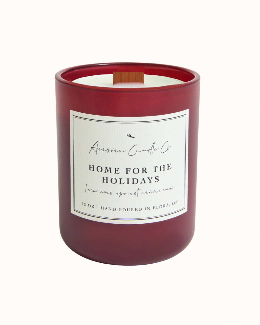 "Home For The Holidays" candle in a deep red coloured jar. There is a white square label on the candle stating the name of the business, scent, "luxe coco apricot crème wax", size, and "hand-poured in Elora, ON".