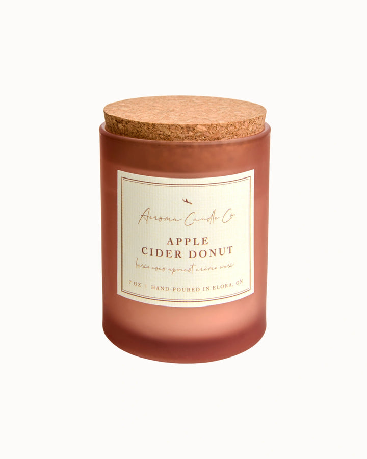 "Apple Cider Donut" candle in an opaque mahogany jar with a cork lid. There is a white square label on the candle stating the name of the business, scent, "luxe coco apricot crème wax", size, and "hand-poured in Elora, ON".