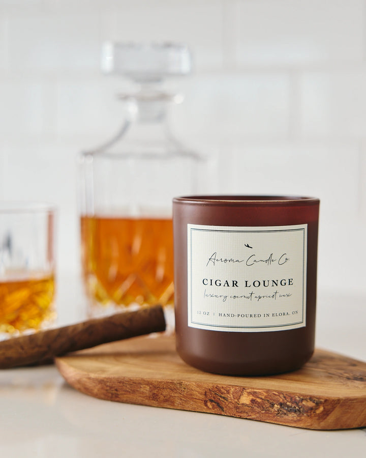 Candle labeled 'Cigar Lounge' on a wooden board with blurred glasses in the background.