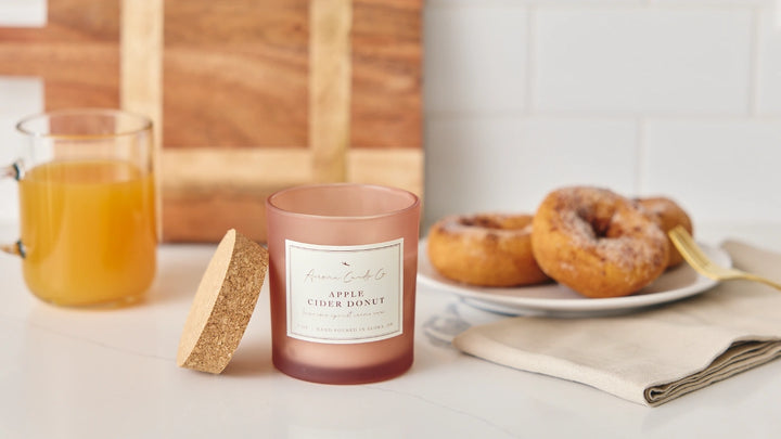 Candle labeled 'Apple Cider Donut' on a table with a plate of donuts and a glass of cider.