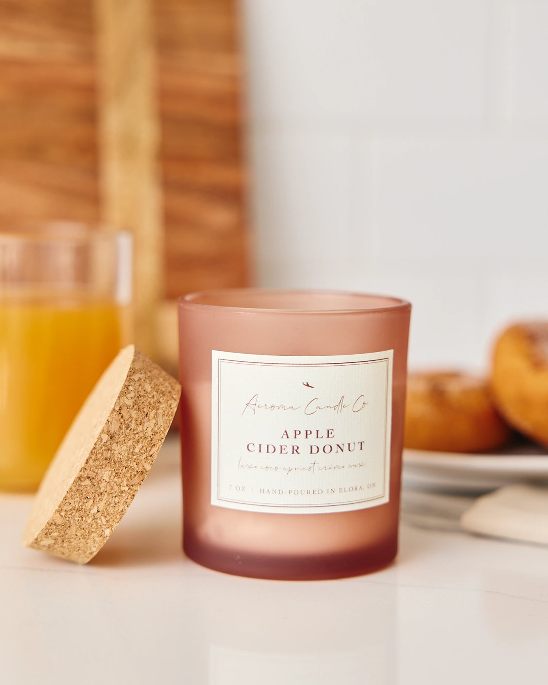 Candle labeled 'Apple Cider Donut' next to a glass of cider and donuts on a wooden surface.