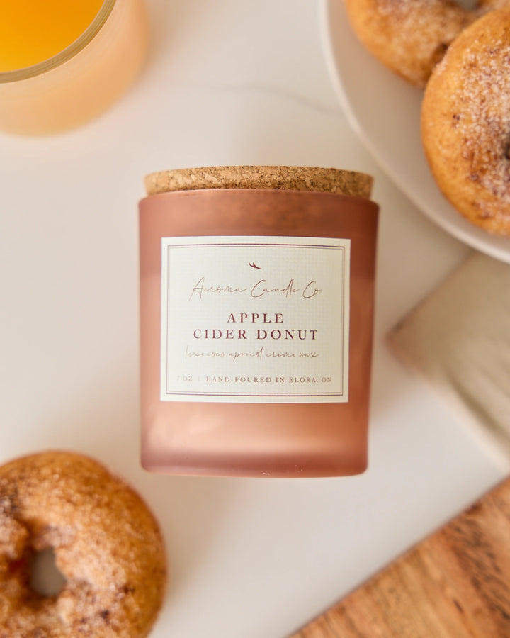 Candle labeled 'Apple Cider Donut' with donuts and a glass of cider on a table.