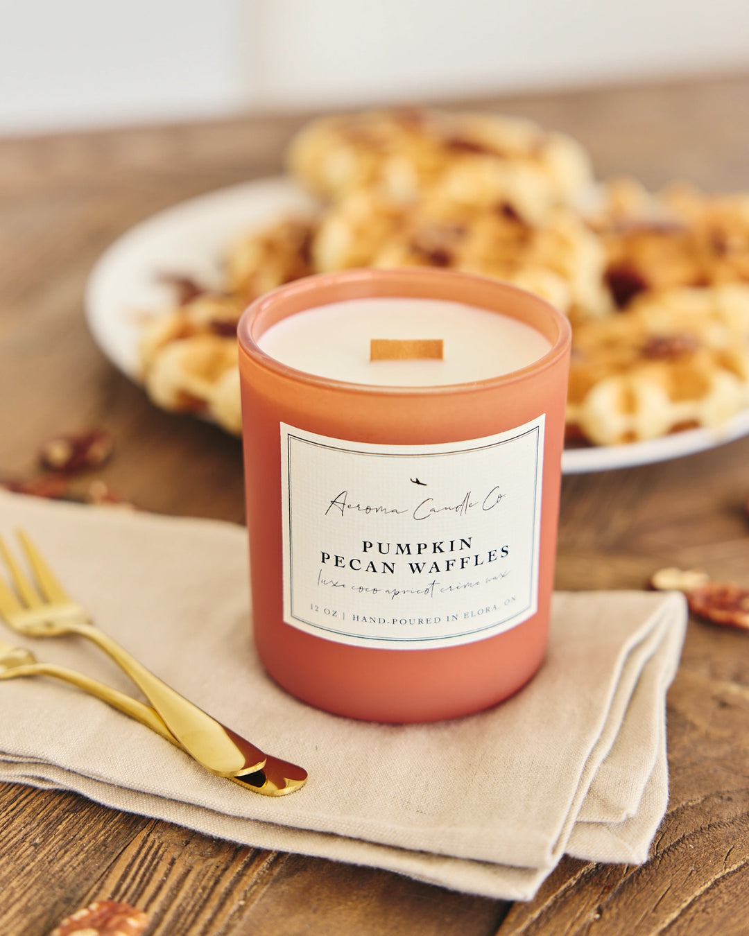 Candle labeled 'Pumpkin Pecan Waffles' on a wooden table with pastries in the background