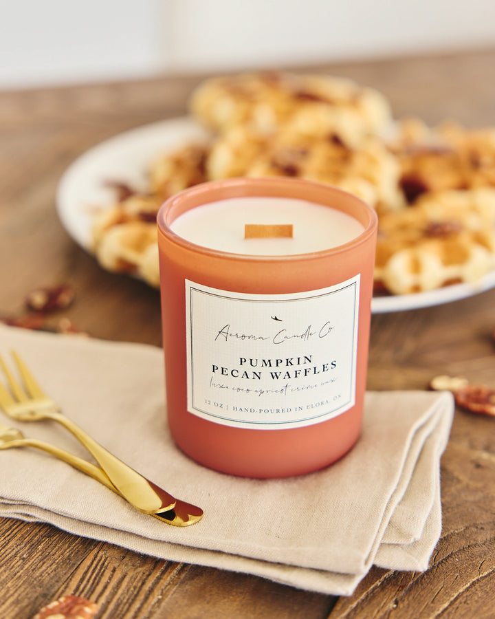 Candle labeled 'Pumpkin Pecan Waffles' on a wooden table with pastries in the background