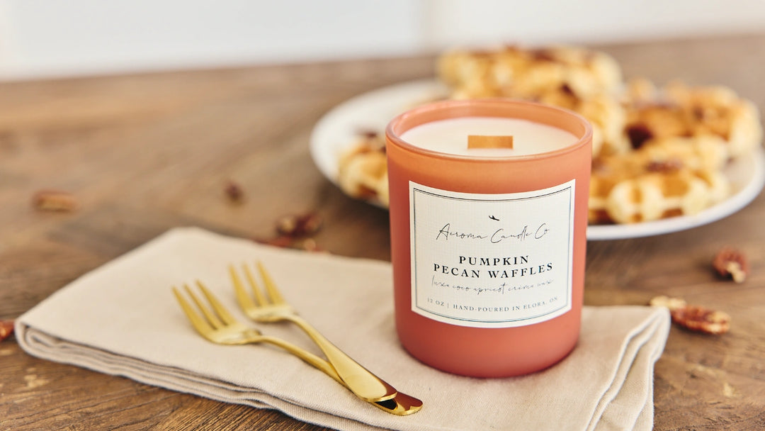 Pink candle labeled 'Pumpkin Pecan Waffles' on a wooden table with waffles and gold forks.
