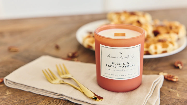 Pink candle labeled 'Pumpkin Pecan Waffles' on a wooden table with waffles and gold forks.