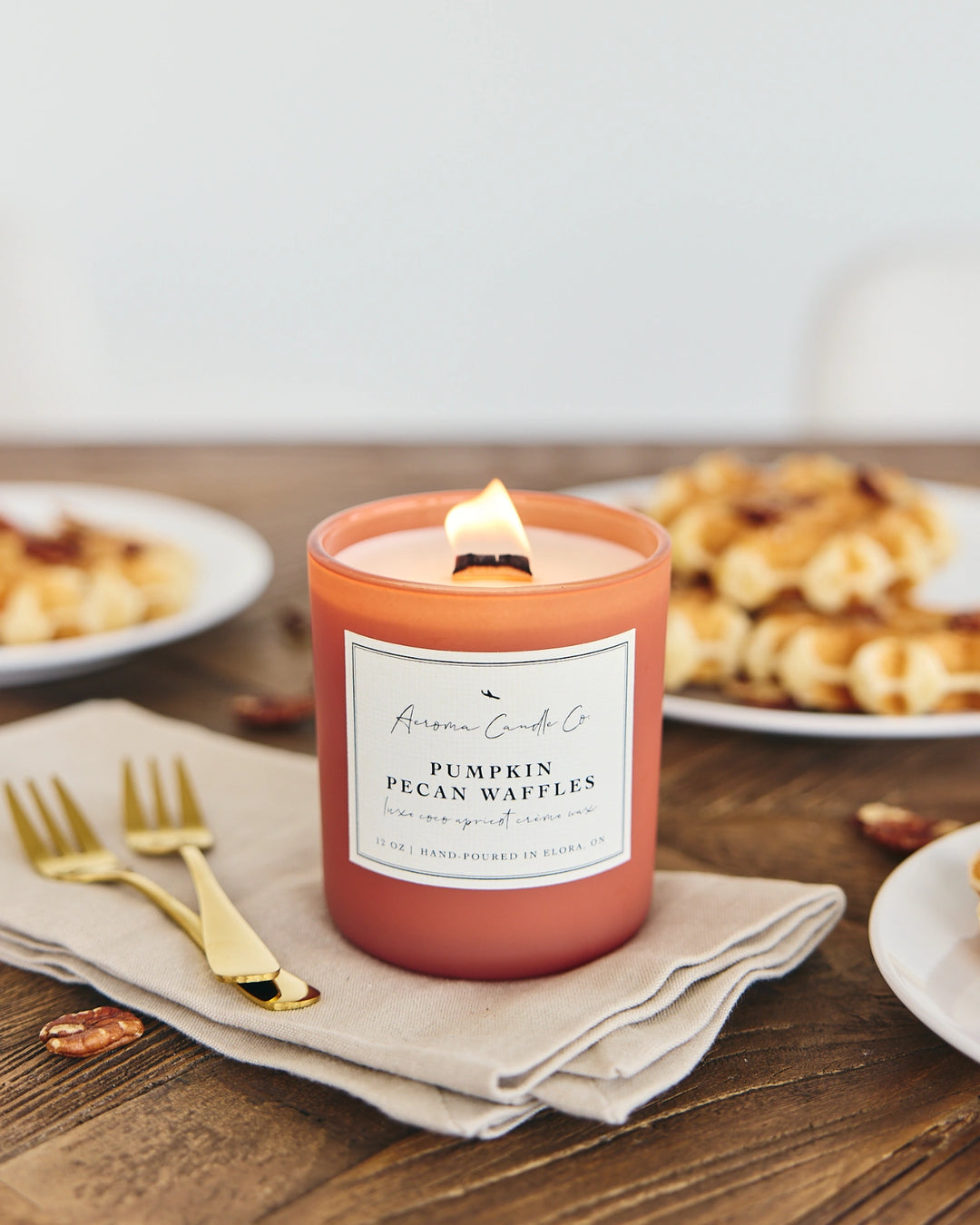 Candle labeled 'Pumpkin Pecan Waffles' on a wooden table with waffles in the background