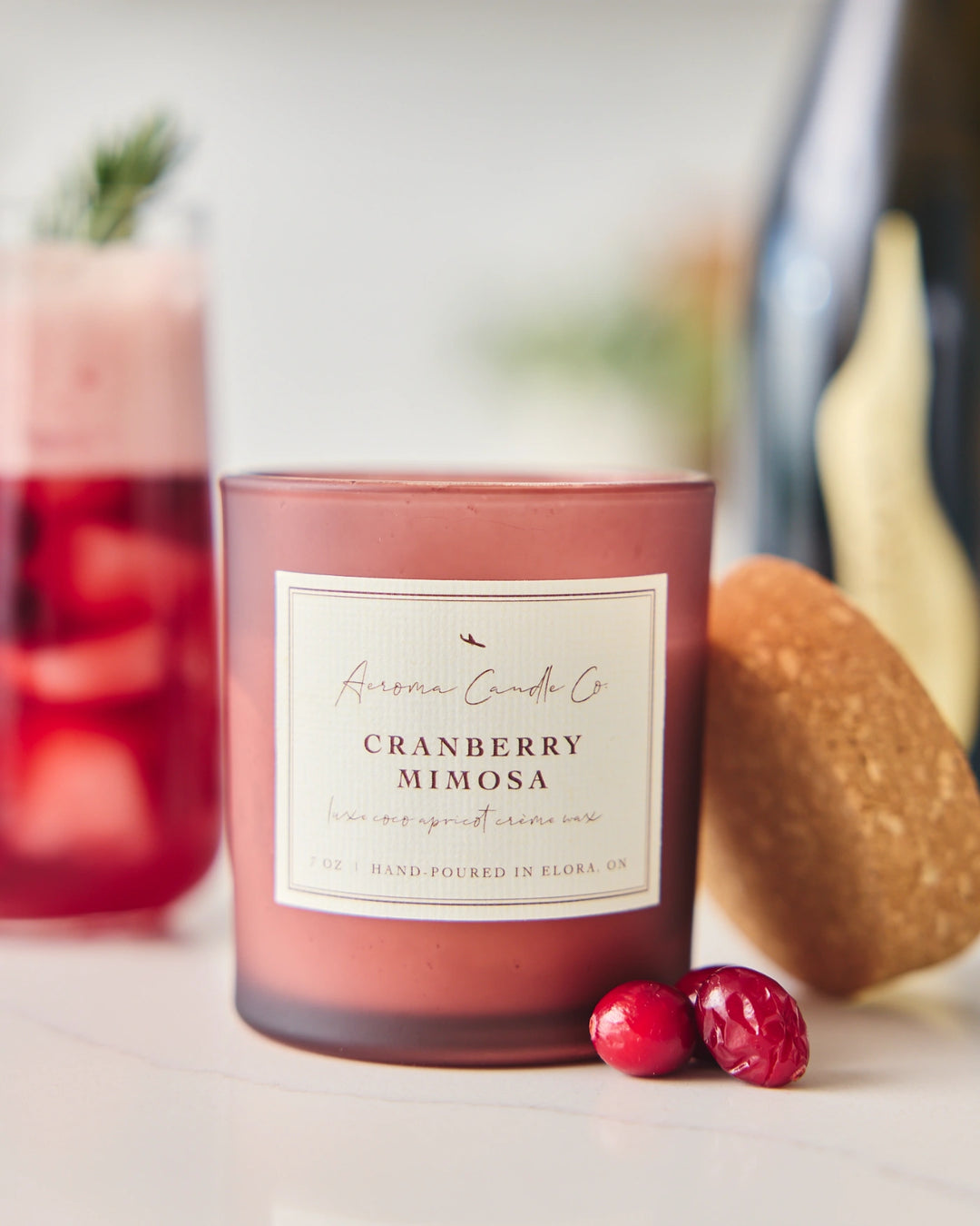 Cranberry Mimosa candle with a glass of cranberry juice and a mimosa in the background.