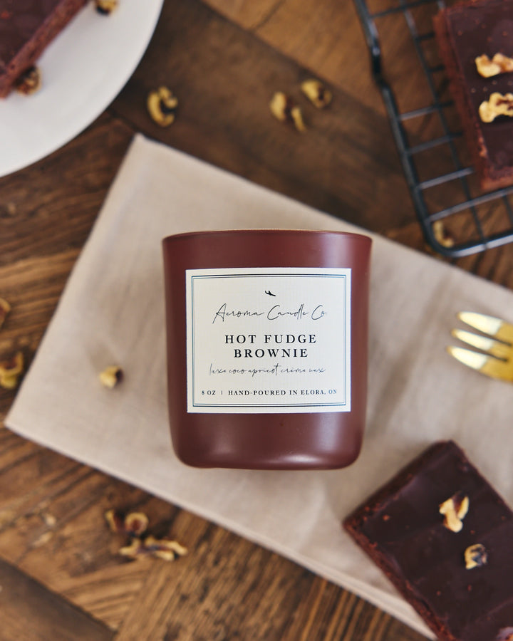 Hot fudge brownie candle with a label on a wooden surface with brownies and nuts.