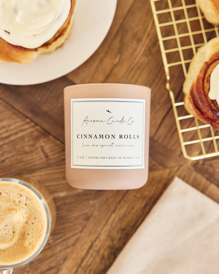Candle labeled 'Cinnamon Rolls' on a wooden surface with cinnamon rolls and coffee.