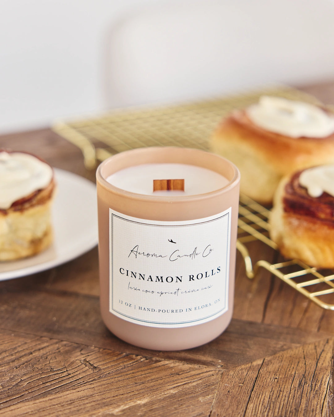 Cinnamon roll-scented candle with cinnamon sticks on a wooden surface with cinnamon rolls in the background.