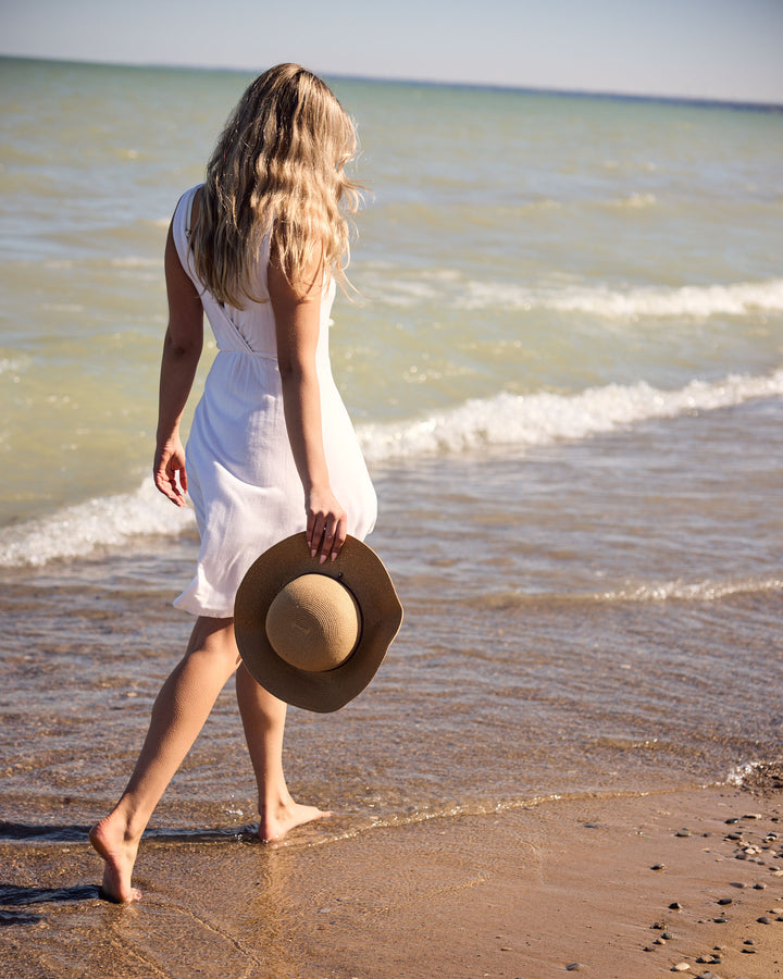 Woman in a white dress holding a hat, walking on a beach.