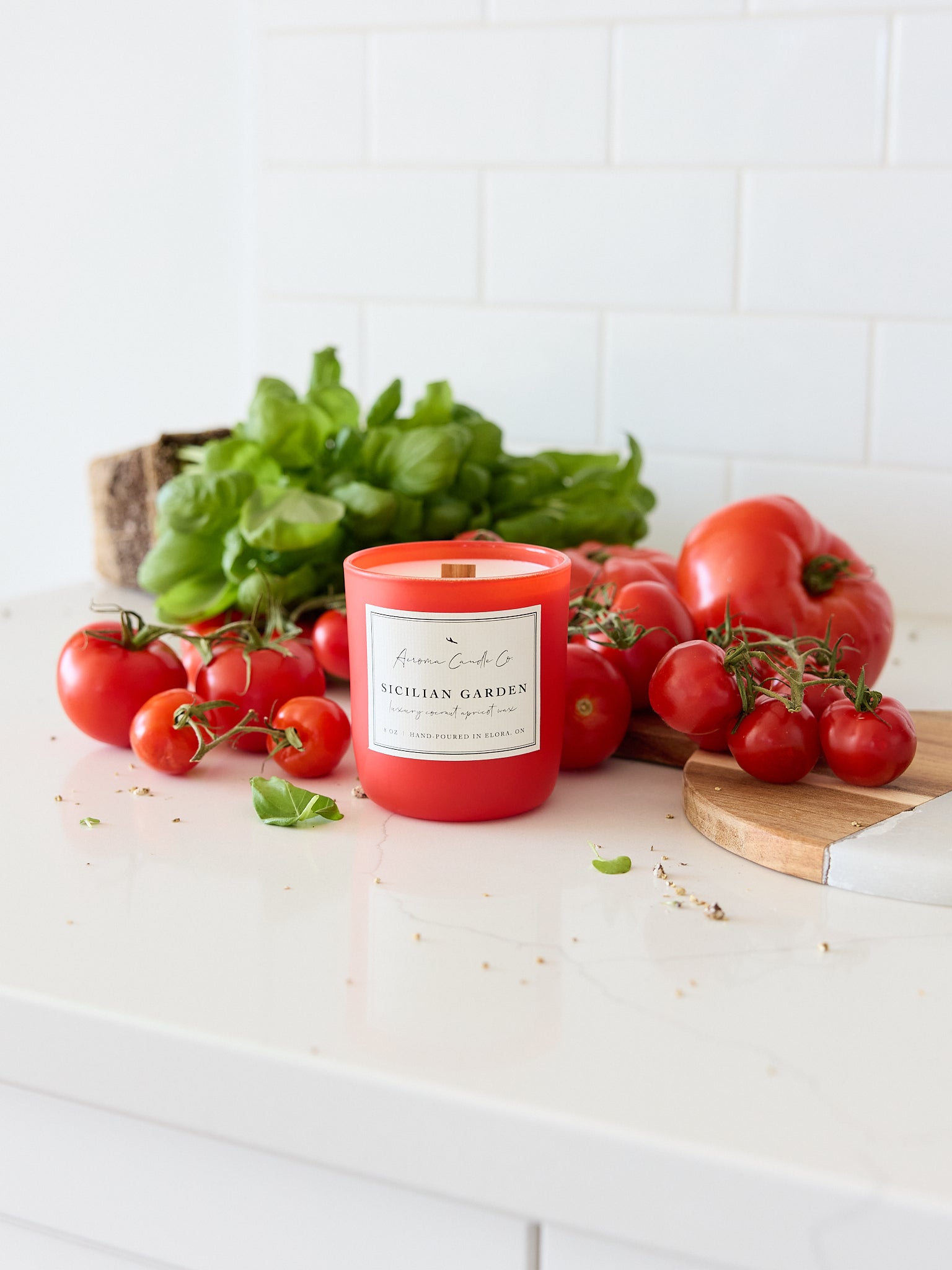 Candle labeled 'Sicilian Garden' surrounded by fresh tomatoes and basil on a white surface.