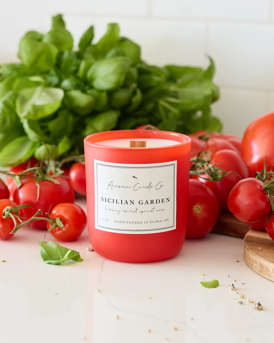 Red candle labeled 'Sicilian Garden' surrounded by fresh tomatoes and basil on a white surface.
