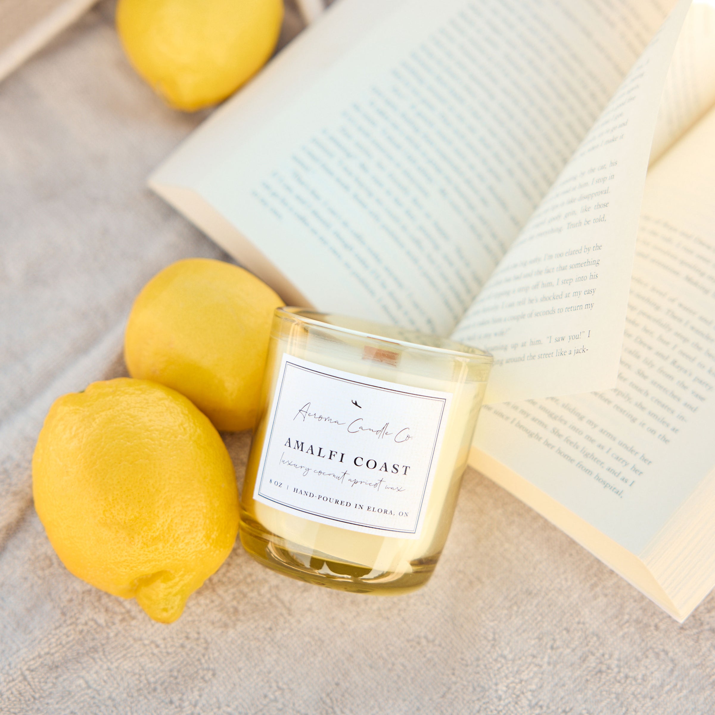 Candle labeled 'Amalfi Coast' with lemons and an open book on a light surface