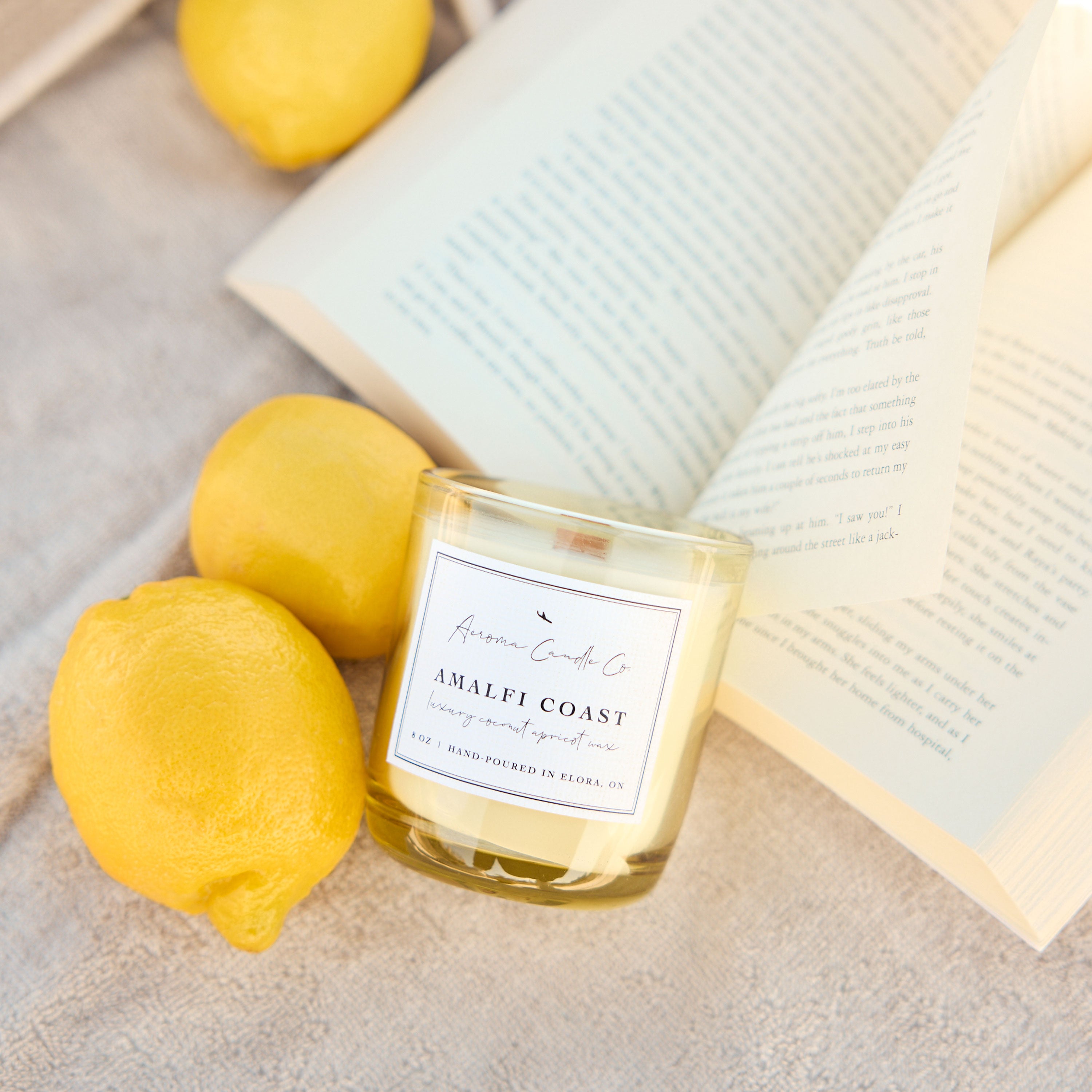 Candle labeled 'Amalfi Coast' with lemons and an open book on a light surface