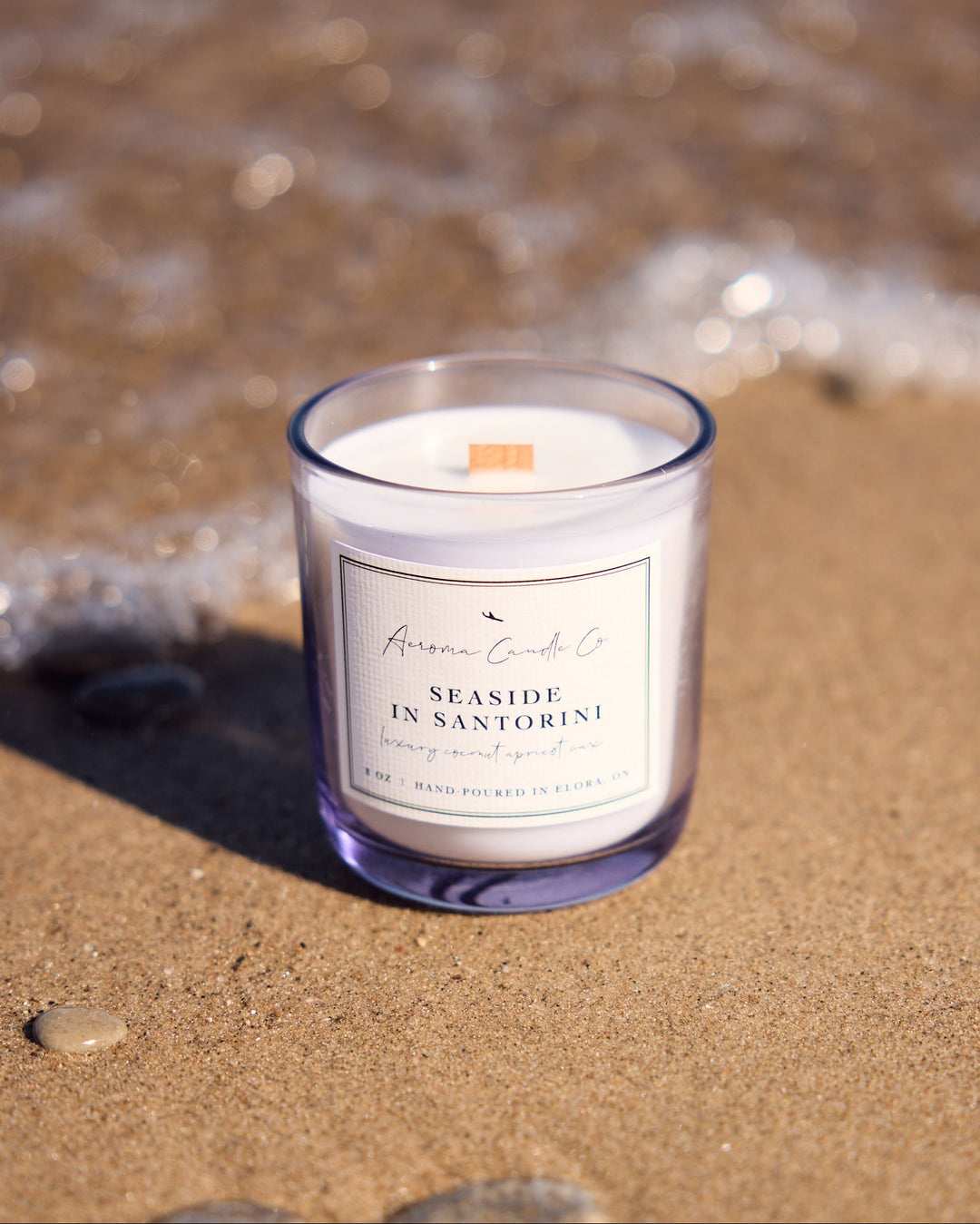 Candle in a glass jar labeled 'Seaside in Santorini' on a sandy beach with waves in the background.