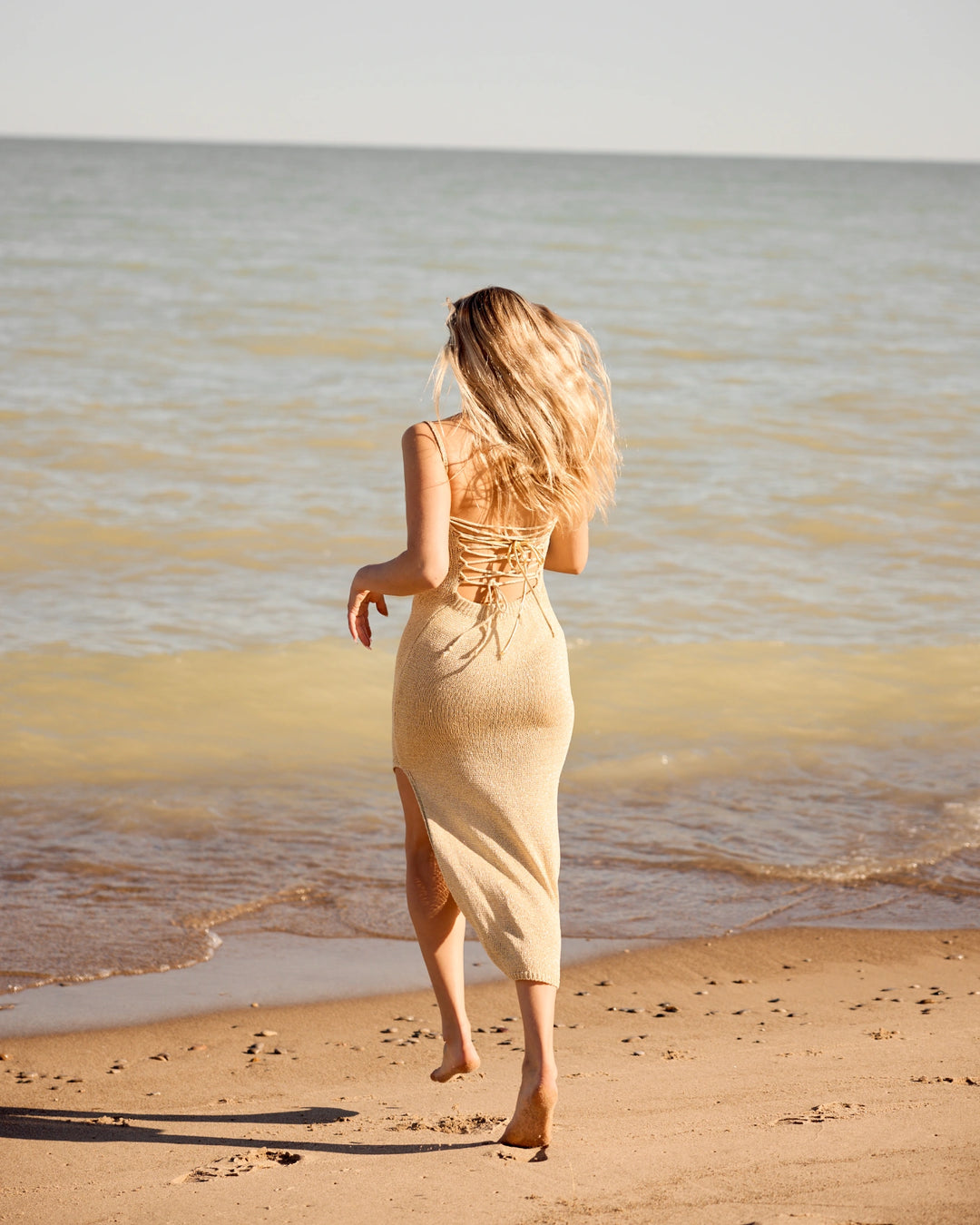 Woman in a beige dress standing on a beach with ocean waves.