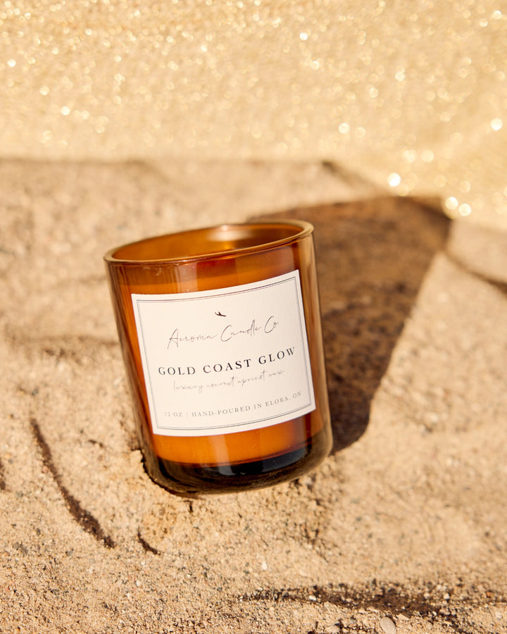 Candle labeled 'Gold Coast Glow' on a sandy surface with a sparkly background