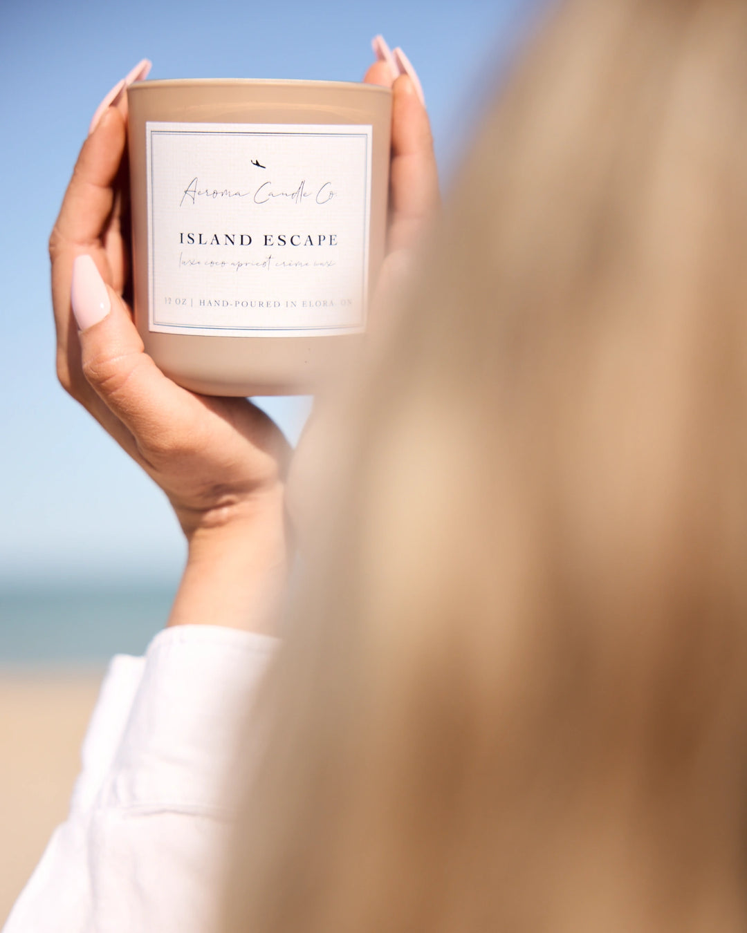 Person holding a 'Island Escape' candle by the beach