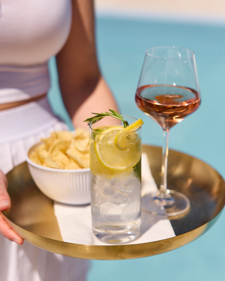 Tray with a glass of water, lemon slice, rose wine glass, and bowl of chips against a blue background