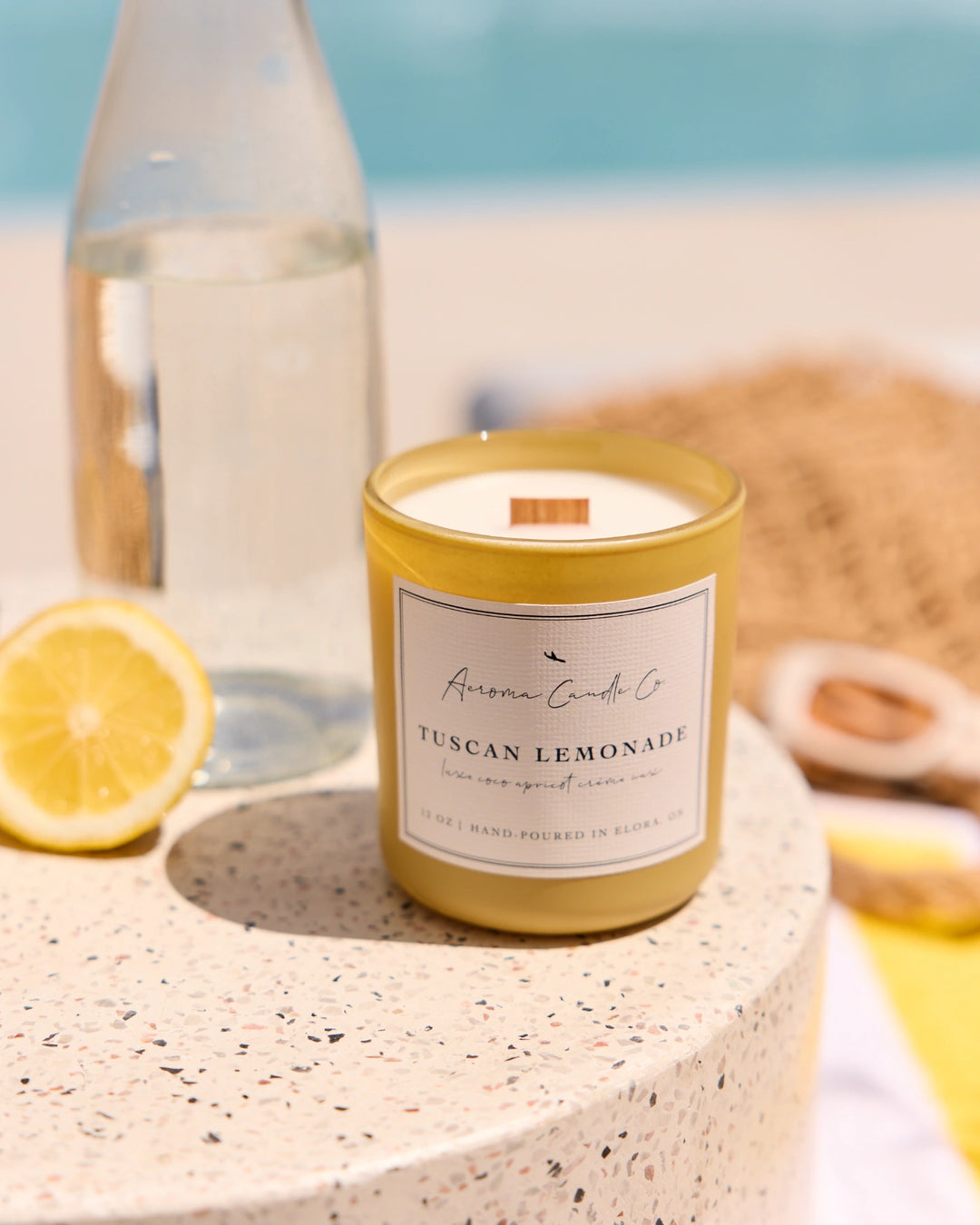 Candle labeled 'Tuscan Lemonade' on a stone surface with lemons and a bottle in the background.