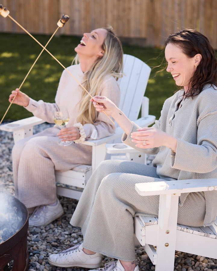 Two women sitting in Adirondack chairs by a fire pit, roasting marshmallows.