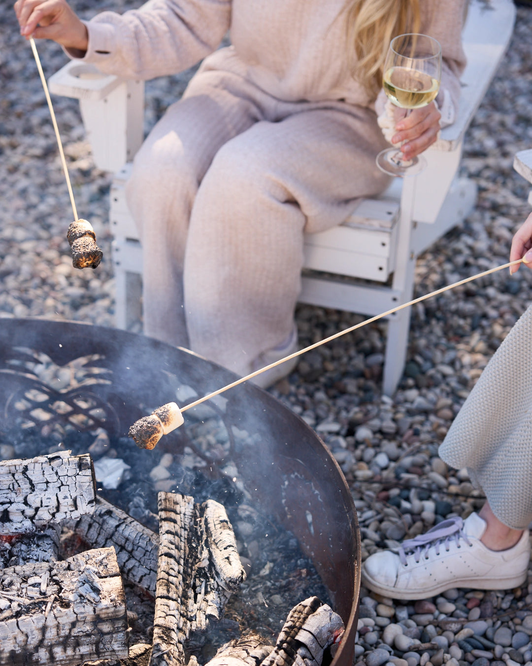 Person roasting marshmallows over a fire pit on a pebbly beach.