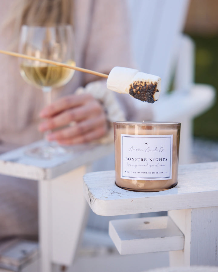 Candle labeled 'Bonfire Nights' with a marshmallow on a stick over it, next to a glass of white wine.